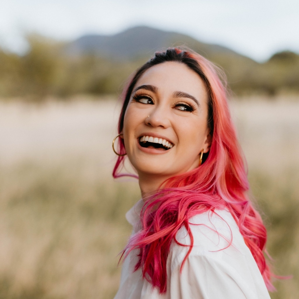 In the foreground, author Erin Baldwin looks back, past the camera, caught mid-laugh. Her hair, pink with black roots, is partially draped over her left shoulder. There's a gold stud in her nose and gold hoops in her ears. The background is a blurry wheat field, a line of trees, and distant mountains against a light gray sky.